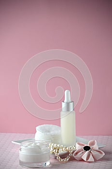 A jar of luxury beauty face cream and serum bottle with pearls on pink color background with copy space.