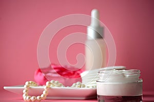 A jar of luxury beauty face cream and serum bottle with pearls on pink color background with copy space.