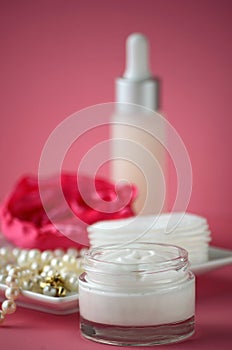 A jar of luxury beauty face cream and serum bottle with pearls on pink color background with copy space.