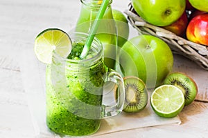 Jar mug with fresh green vegetable smoothie, bottle with fruit juice, apples in basket, lime, kiwi,bright sun fleck