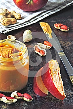 Jar of peanut butter, peanuts in a peel, apple slices and a knife.