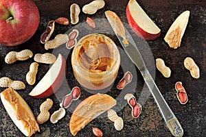 Jar of peanut butter, peanuts in a peel, apple slices and a knife.