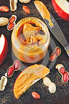 Jar of peanut butter, peanuts in a peel, apple slices and a knife.