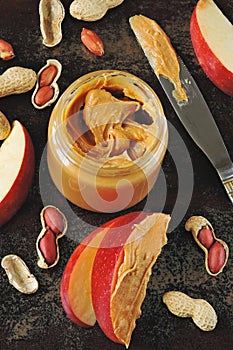 Jar of peanut butter, peanuts in a peel, apple slices and a knife.