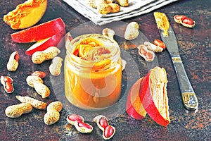 Jar of peanut butter, peanuts in a peel, apple slices and a knife.