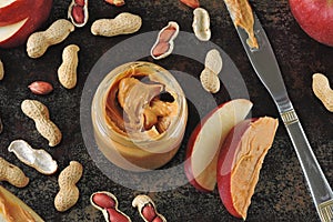 Jar of peanut butter, peanuts in a peel, apple slices and a knife.