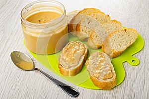 Jar with peanut butter, slices of bread, sandwiches with peanut paste on cutting board, spoon on wooden table