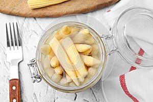 Jar of pickled baby corn and fork on white textured table, flat lay