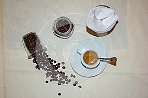 Jar with spilled coffee beans on linen tablecloth with cup of espresso full with spoon.