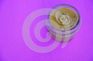 Jar and spoon of peanut butter and peanuts on purple background from top view. copy space