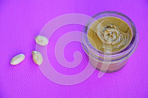 Jar and spoon of peanut butter and peanuts on purple background from top view. copy space