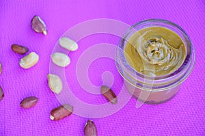 Jar and spoon of peanut butter and peanuts on purple background from top view. copy space