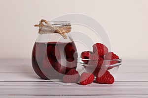 Jar of tasty canned raspberry jam and fresh berries in glass bowl on white wooden table