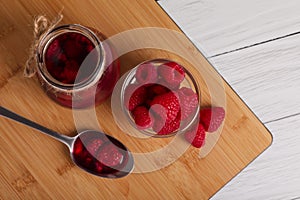 Jar of tasty canned raspberry jam and fresh berries in glass bowl on white wooden table, flat lay