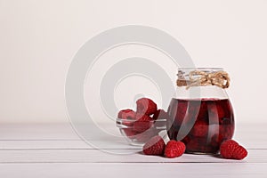 Jar of tasty canned raspberry jam and fresh berries in glass bowl on white wooden table, space for text