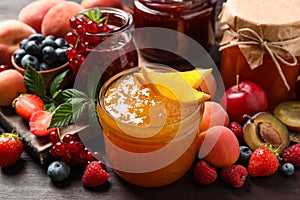 Jars with different jams and fresh fruits on wooden table, closeup