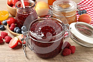 Jars with different jams and fresh fruits on wooden table, closeup