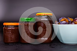 Jars with plum jam on a black background. In the background bowls with plums