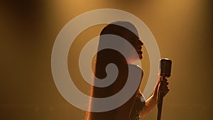 Jazz vocalist in glowing elegant dress perform on stage. Side view of a silhouette of an attractive woman singing into a