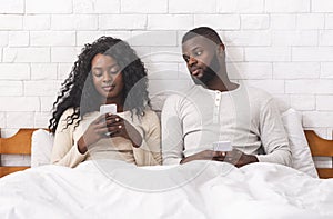 Jealous man peeking into wife`s cellphone sitting on bed together