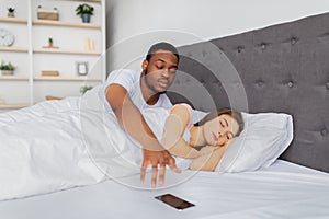 Jealous Afro man checking his sleeping girlfriend& x27;s mobile phone in bed at home. Marriage problems concept