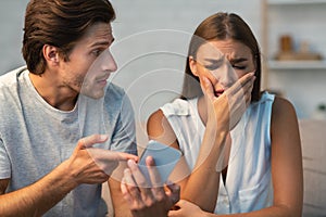Jealous Boyfriend Showing Phone To Girlfriend Sitting On Couch Indoor