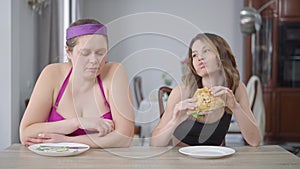 Jealous fat woman looking at slim Caucasian friend eating sandwich. Front view of slender and obese women sitting at