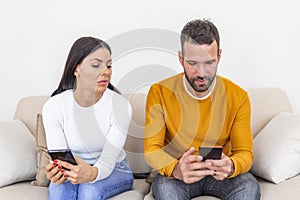 Jealous young woman with smartphone looking at smiling boyfriend using smartphone at home, relationship problem concept. Mistrust