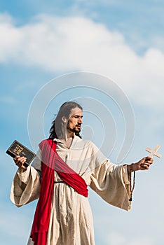 Jesus holding holy bible and wooden cross against blue sky