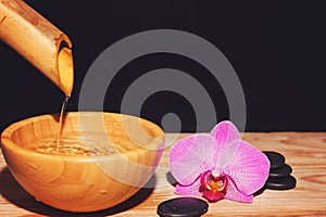 The jet of water is poured from the bamboo into a bowl on a wooden table, next to the spa treatment stones and the orchid flower,