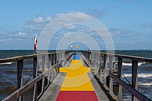 A jetty with a colourful carpet in blue, yellow and red. A blue ocean and sky in the background