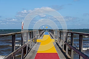 A jetty with a colourful carpet in blue, yellow and red. A blue ocean and sky in the background