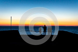 A jetty or pier with silhouetted people with a sunset at sea.