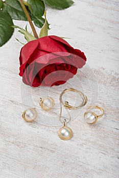 Jewelry set of golden ring, earrings, necklace with pearls and red rose on white wooden background