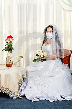 A Jewish bride sits in a synagogue before a chuppa ceremony during a pandemic wearing a medical mask and a bouquet of