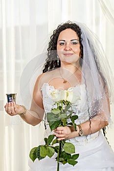 A Jewish bride stands in the hall before the chuppa ceremony with a bouquet of white roses in her hands and a glass of wine