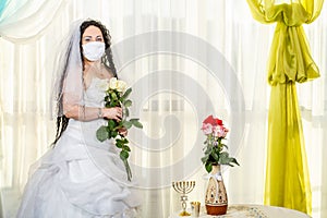 A Jewish bride in a synagogue at a table with flowers before a chuppa ceremony during a pandemic, wearing a medical mask