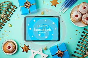 Jewish holiday Hanukkah concept with digital tablet mock up, menorah, sufganiyah and gift box over blue background