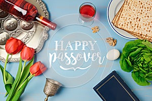 Jewish holiday Passover Pesah greeting card with seder plate, matzoh, tulip flowers and wine bottle on wooden background.