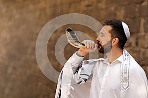 Jewish man blowing shofar on Rosh Hashanah outdoors. Wearing tallit with words Blessed Are You, Lord Our God, King Of The Universe