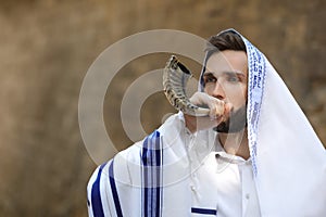 Jewish man blowing shofar on Rosh Hashanah outdoors. Wearing tallit with words Blessed Are You, Lord Our God, King Of The Universe