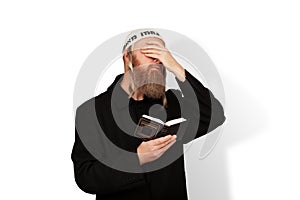 Jewish man holding Siddur covering eyes with hand on white background. Bearded Jew hasid with sidelocks in white kippah