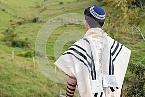 Jewish man praying with talit, kippah and tefillin in nature with beautiful green meadow in the background.