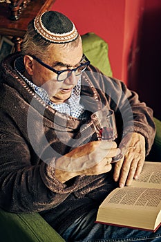 Jewish senior in the armchair reading a torah book and drinking kosher wine