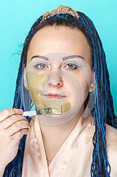 Jewish woman with Afro braids applies a mask of green clay to her face with a brush on a blue background.