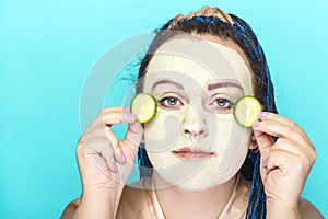 Jewish woman with blue Afro braids face in a frozen mask of green clay with pieces of cucumber in her hands.