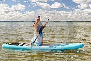 A Jewish woman in a headscarf and a pareo on her knees on a SUP board with an oar swims in a lake on a sunny day.