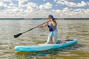 A Jewish woman in a kerchief kisuy rosh in a pareo on her knees on a SUP board swims in the lake on a sunny day.