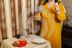 A Jewish woman in a wig and head covering at home at the table at the Passover seder with an egg and a jug of salt water