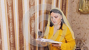 A Jewish woman in a wig and head covering makes a celebratory kdush with a glass in her hand and a siddur.